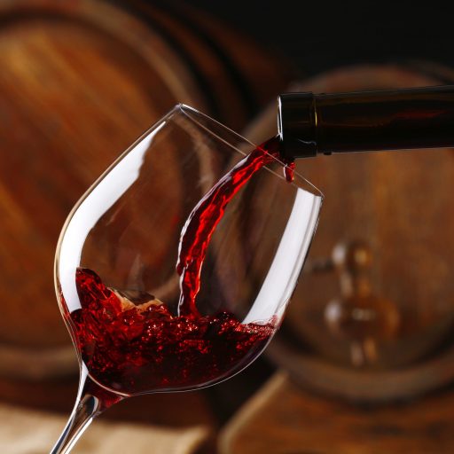 Pouring red wine from bottle into glass with wooden wine casks on background